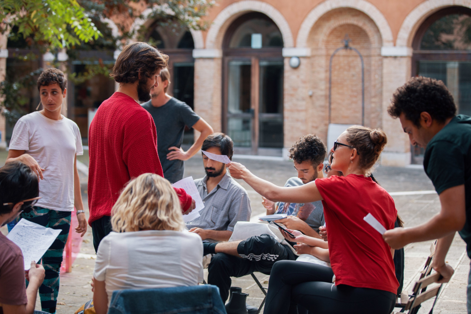 A group of young adults takes part in an outdoor activity in a courtyard with brick arches. Some are reading or holding papers, others interact with one another, while a blindfolded participant receives a piece of paper from someone. The setting feels collaborative and informal.