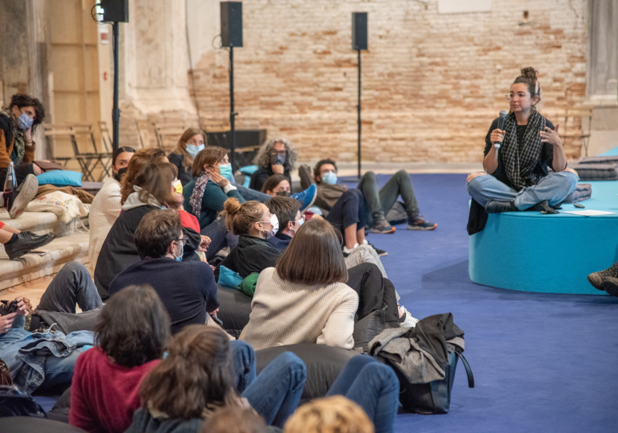 A speaker sits cross legged on a blue platform holding a microphone and addressing an audience seated informally on the floor and cushions inside a spacious hall with exposed brick walls.