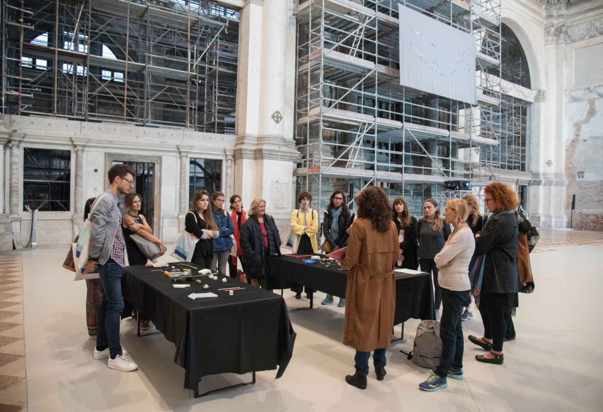 A group of adults gathers around two black tables inside a large historic hall with scaffolding along the walls, listening to a woman who is presenting materials laid out in front of her.