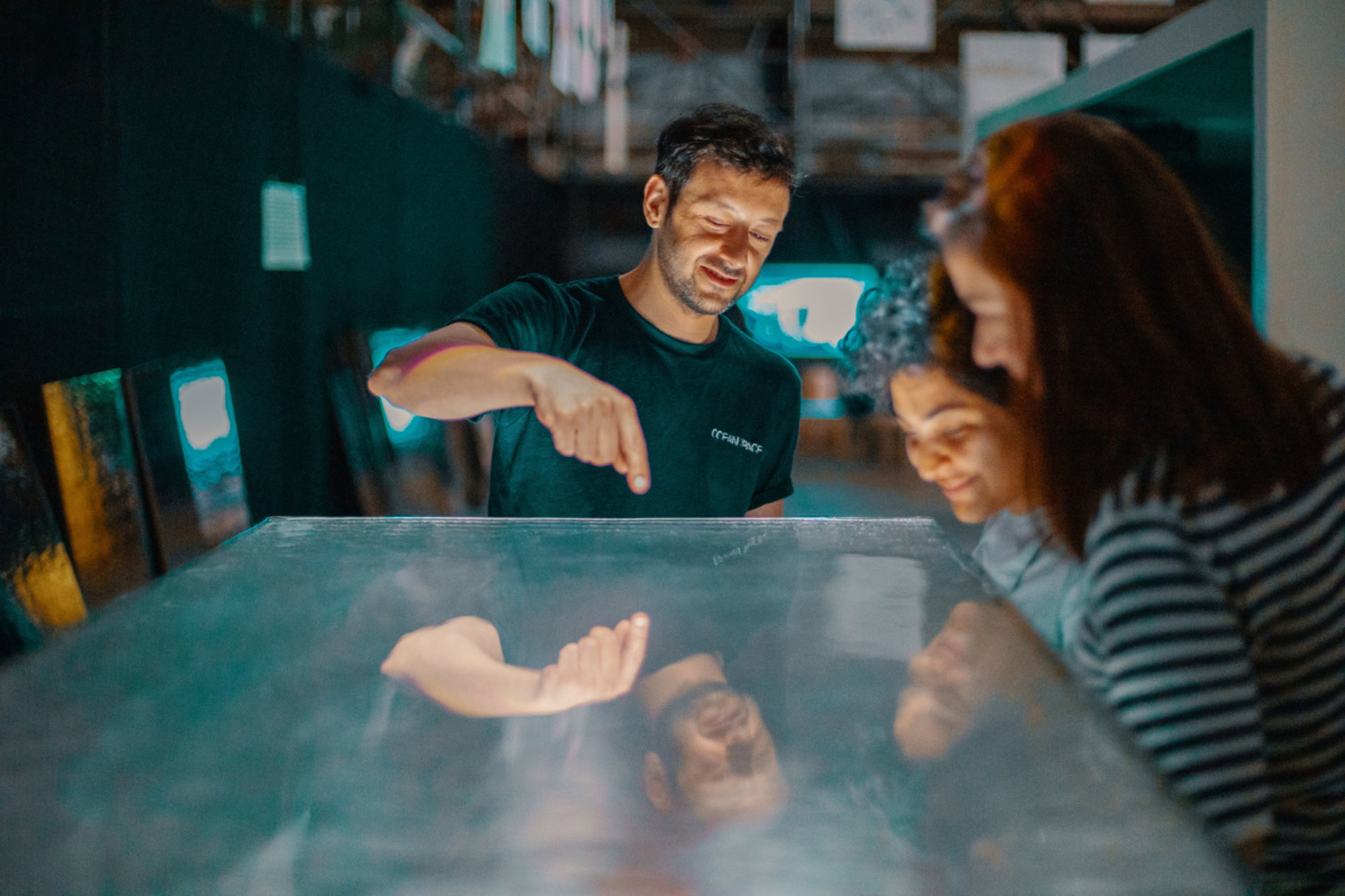 A man points at a glowing interactive surface while two women lean in, watching closely and smiling in a dimly lit, futuristic exhibition space.