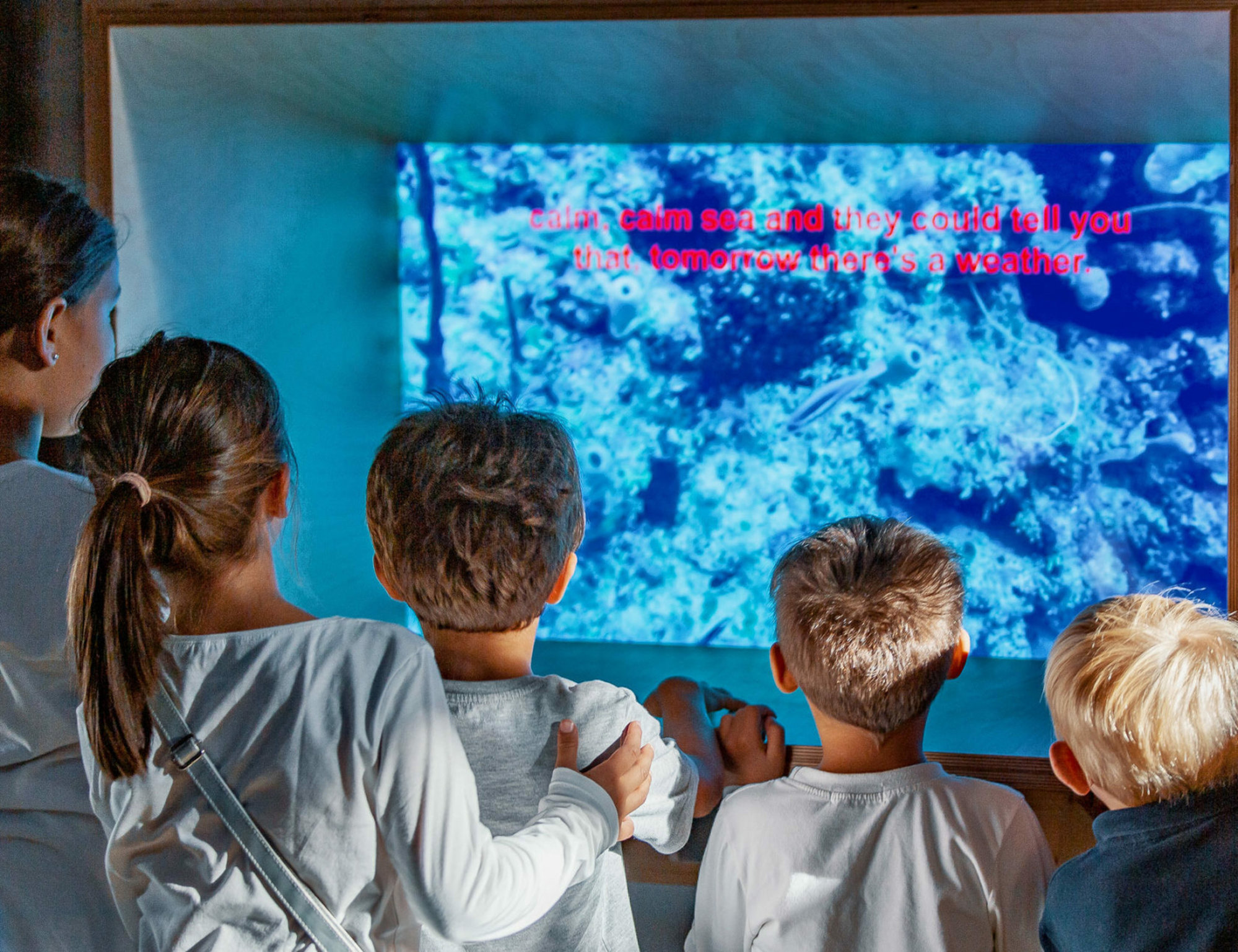 A group of young children stands closely together watching an underwater video projection with red text subtitles, their faces lit by the blue glow of the screen.