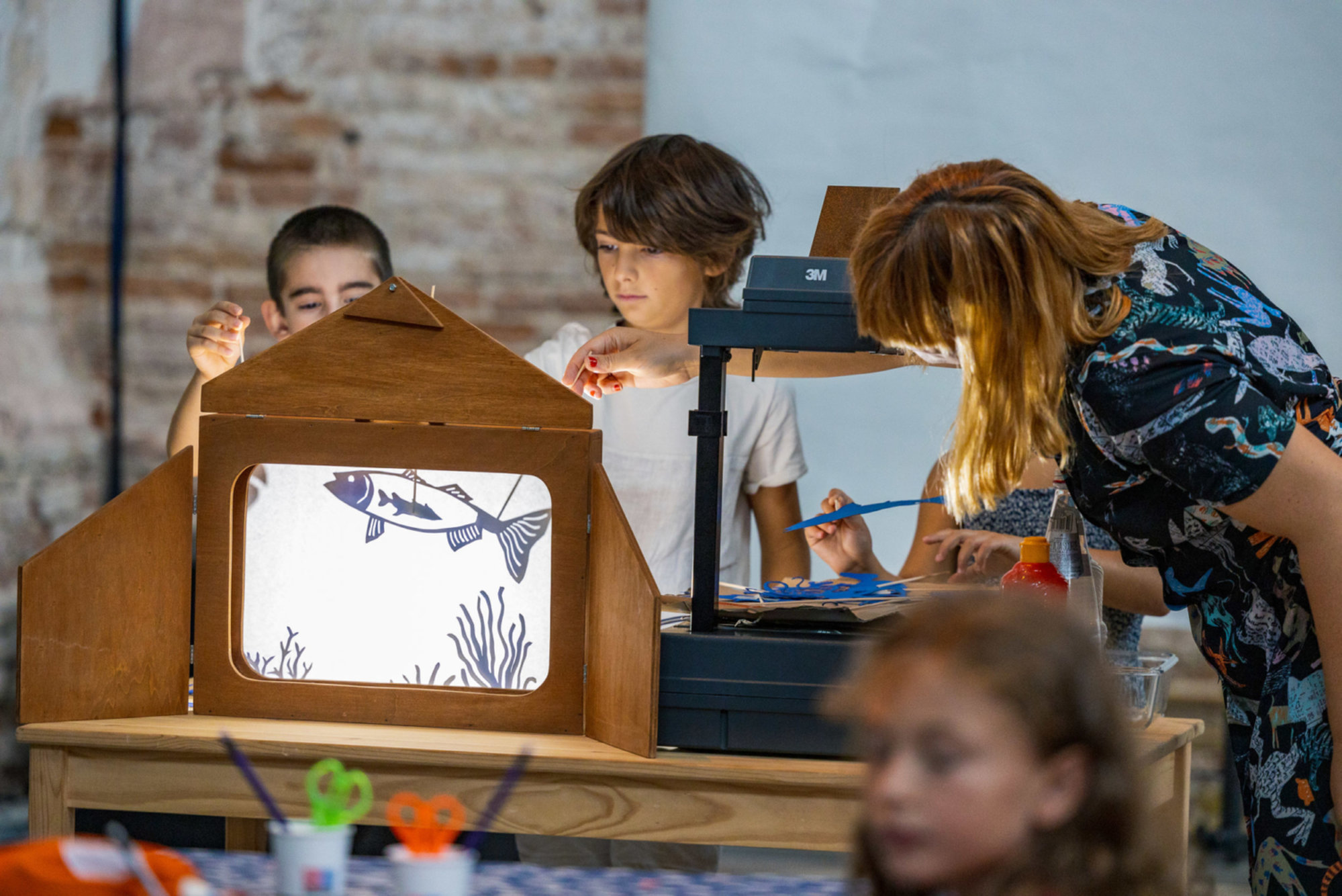Children work with a teacher on a shadow theater setup, using cut-out fish and seaweed shapes illuminated by an overhead projector as they create an underwater scene.