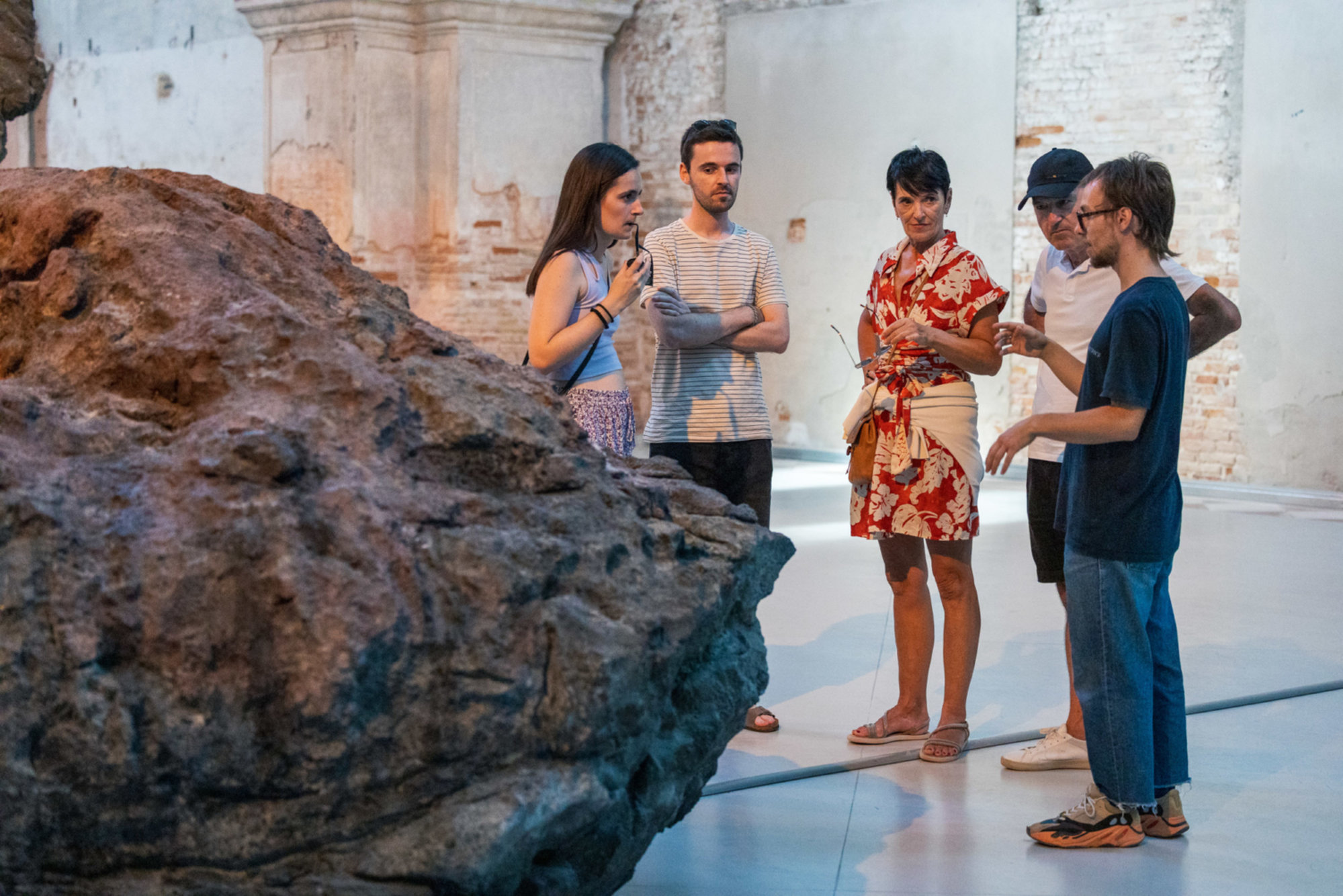 A group of people stands inside a spacious, rustic exhibition hall, listening attentively as a guide explains a large textured rock installation in the foreground.