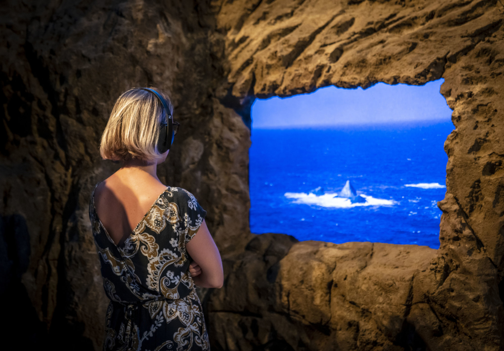 A woman wearing headphones stands inside a dim, cave like installation, watching a bright video projection of the ocean and a breaking wave framed by a rough stone window.