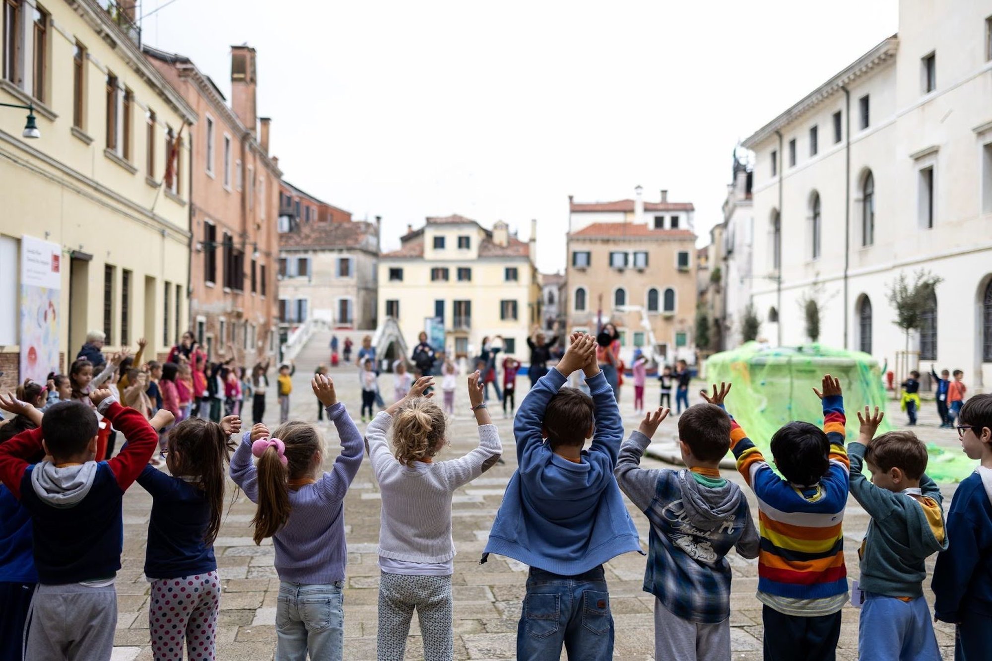 A group of children standing in a circle during an outdoor activity in the square in front of the Church of San Lorenzo in Venice, raising their hands while historic buildings line the background.
